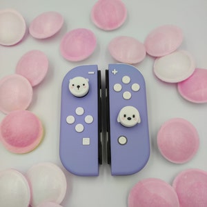Può includere: Un controller Nintendo Switch viola con pulsanti frontali personalizzati che raffigurano un orso polare e una foca. Il controller è circondato da caramelle rosa e bianche.