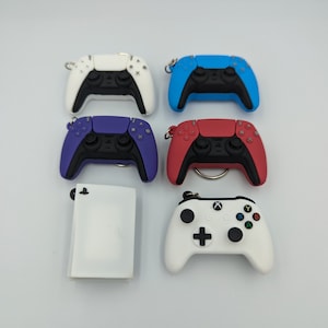Könnte beinhalten: Ein Set mit sechs Schlüsselanhängern, die verschiedene farbige Videospielcontroller zeigen. Die Schlüsselanhänger umfassen einen weißen, blauen, violetten, roten und zwei weißen Controller. Die Controller sind alle Miniaturversionen beliebter Spielkonsolen.