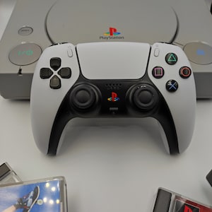 Op de afbeelding: Een witte en zwarte PlayStation 5-controller naast een grijze PlayStation 1-console. De PlayStation 1-console heeft de tekst "PlayStation" op de voorkant.