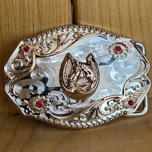 Op de afbeelding: Zilveren en gouden gesp met een hoefijzerontwerp en rode strass steentjes. De gesp heeft een sierlijk, gegraveerd ontwerp.