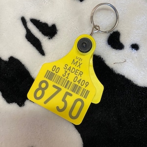 May include: A yellow livestock ear tag with the text "MX SADER" and the number "8750" printed on it. The tag is attached to a silver key ring. The background is a black and white cow print.