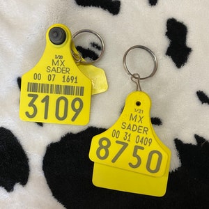 May include: Two yellow livestock ear tags with silver key rings. One tag displays the number 3109, the other displays 8750. Both tags have printed text including the name "MX SADER" and a date.