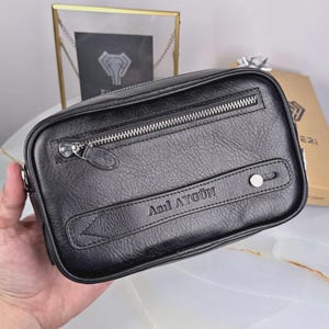 Könnte beinhalten: Eine schwarze, rechteckige Ledertasche mit strukturierter Oberfläche, ausgestattet mit einer horizontalen Reißverschlusstasche oben und einem dekorativen Lederriemen mit silberfarbenem Metallniet. Der Name Anıl AYGÜN ist vorne eingeprägt.