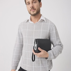 May include: A black leather clutch with a wrist strap. The clutch has a silver zipper and a small silver accent. The person is wearing a gray sweater with a white grid pattern and a white collared shirt underneath.