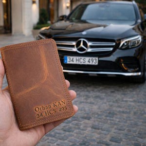 Op de afbeelding: Een bruine leren portemonnee met de tekst "Orhan KAN 34 HCS 493" in reliëf op de voorkant. De portemonnee wordt in een hand gehouden, met een zwarte auto op de achtergrond.