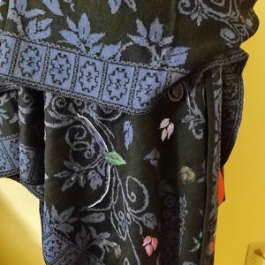 May include: A dark green knitted poncho with a floral pattern in shades of blue, gray, and white. The poncho features a blue border with a geometric design and embroidered leaves in various colors.