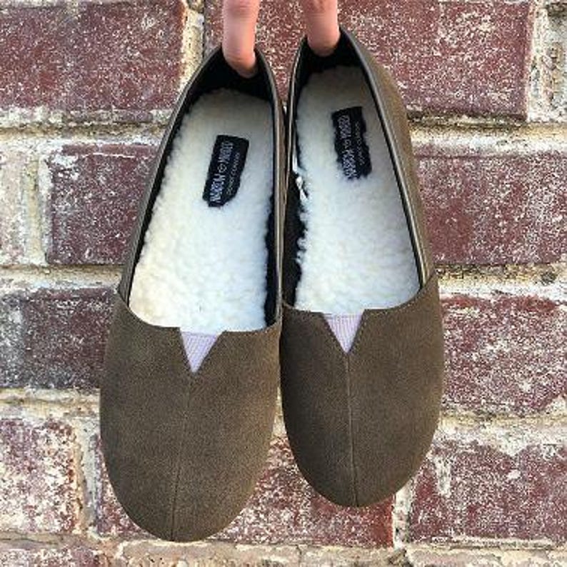 K&ouml;nnte beinhalten: Ein Paar olivgr&uuml;ne Slipper aus Wildleder mit einem Futter aus Kunstfell. Die Schuhe haben eine spitze Zehenpartie und eine flache Sohle.