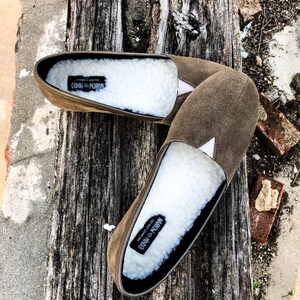 K&ouml;nnte beinhalten: Ein Paar braune Wildlederslipper mit wei&szlig;em Kunstfellfutter. Die Slipper haben ein schwarzes Etikett auf der Innenseite mit der Aufschrift "Narrow Minded Do Not Conform".