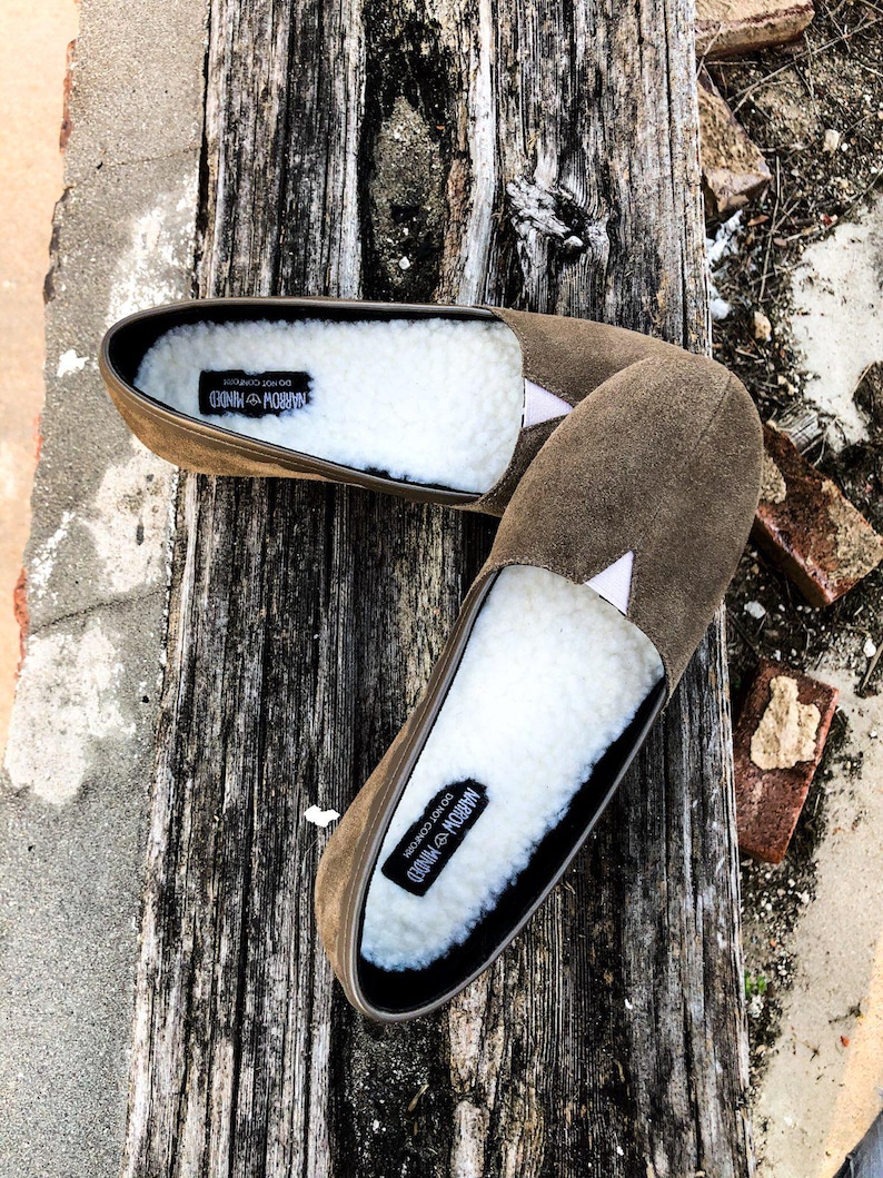 K&ouml;nnte beinhalten: Ein Paar braune Wildlederslipper mit wei&szlig;em Kunstfellfutter. Die Slipper haben ein schwarzes Etikett auf der Innenseite mit der Aufschrift "Narrow Minded Do Not Conform".