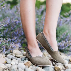 K&ouml;nnte beinhalten: Ein Paar graue Wildlederschuhe mit gewellten Kanten und einem dekorativen Stichdetail. Die Schuhe werden von einer Person getragen, die auf einem Bett aus lila Blumen steht.