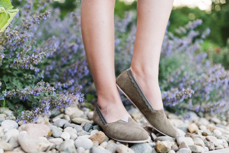 K&ouml;nnte beinhalten: Ein Paar graue Wildlederschuhe mit gewellten Kanten und einem dekorativen Stichdetail. Die Schuhe werden von einer Person getragen, die auf einem Bett aus lila Blumen steht.