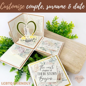 May include: A white and gold explosion box with a floral design. The box has a pop-up feature with a couple standing in front of a heart. The box also has a label that says "Mr & Mrs Manley" and a date that says "8th August 2023". The box is decorated with flowers, leaves, and a gold ribbon. The text "The next chapter of THEIR STORY Begins..." is on one of the panels.