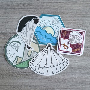 May include: Five colorful stickers featuring paragliding designs. The stickers are round, hexagonal, and square. The stickers feature black and white illustrations of paragliders, mountains, and clouds. The stickers have text that reads "SOAR", "CROSS COUNTRY", and "HANDLESKIES.COM".