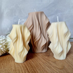 May include: Three decorative candles with a unique, draped design. Two are a creamy off-white, and one is a light tan. The candles are on a wooden surface with a white flower and a light gray background.