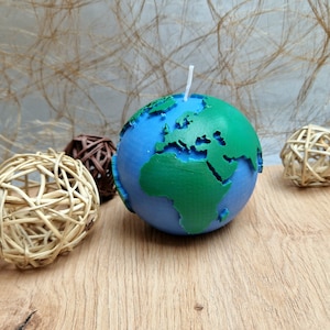 Pode incluir: Uma vela azul e verde em forma de Terra com um pavio branco. A vela tem continentes verdes e oceanos azuis. Esferas decorativas tecidas em tons de castanho e bege são dispostas ao redor da vela sobre uma superfície de madeira.