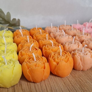 Könnte beinhalten: Sortiment kleiner, dekorativer blumenförmiger Kerzen in Gelb, Orange und Rosa. Die Kerzen sind in Reihen auf einer Holzoberfläche angeordnet.