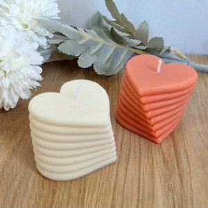 Pode incluir: Duas velas em forma de coração, uma branca e outra laranja, estão empilhadas em camadas. As velas estão sobre uma superfície de madeira.