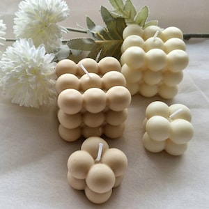 Könnte beinhalten: Vier beige und weiße Blasenkerzen in verschiedenen Größen. Die Kerzen sind quadratisch und rund geformt und haben eine strukturierte Oberfläche. Die Kerzen sind auf einer weißen Oberfläche angeordnet.