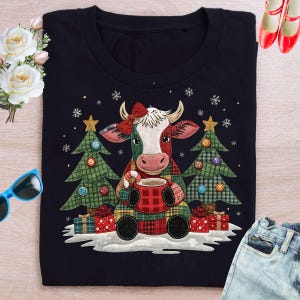 Puede incluir: Camiseta negra con un diseño de vaca navideña. La vaca lleva un lazo rojo y sostiene una taza, flanqueada por árboles de Navidad y regalos. Los copos de nieve y las estrellas completan el tema festivo.