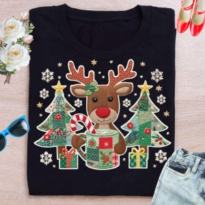 Puede incluir: Camiseta negra de cuello redondo con un diseño de reno con nariz roja, sosteniendo una taza y un bastón de caramelo. Dos árboles de Navidad de patchwork flanquean el reno. El diseño incluye copos de nieve y estrellas.