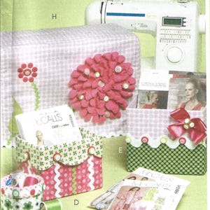 May include: A sewing machine with a white body and digital display is featured. Accessories include fabric storage boxes, sewing patterns, and a decorative flower. The boxes have various patterns and colors, including pink, green, and white.