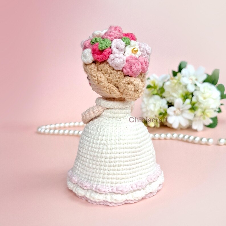以下が含まれることがあります： 白いドレスにピンクのフリルと花冠をつけた、かぎ針編みの人形。人形は茶色の髪で、ピンクの表面に立っています。