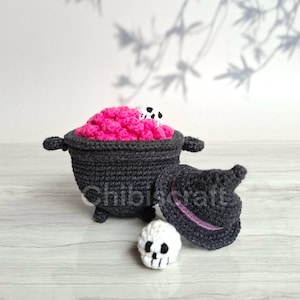 May include: A crocheted cauldron with a pink, textured filling and a small white skull. A matching witch's hat and a separate skull are also present. The cauldron is dark gray, with the text "Chibiscraft" visible.