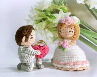 以下が含まれることがあります： かぎ針編みで作られた結婚式の人形。新郎はひざまずいて、ハート型の箱に2つの金の指輪を持っています。花嫁はピンクのリボンと花冠をつけた白いドレスを着ています。