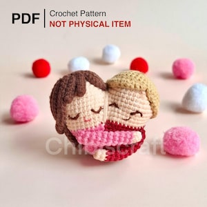 Könnte beinhalten: Eine herzförmige Häkelpuppe mit zwei Figuren, die sich umarmen, mit braunen und blonden Haaren und Kleidung in Rosa und Rot. Der Text "PDF Crochet Pattern NOT PHYSICAL ITEM" steht oben. Kleine, flauschige Pompons in Rot, Rosa und Weiß sind verstreut.