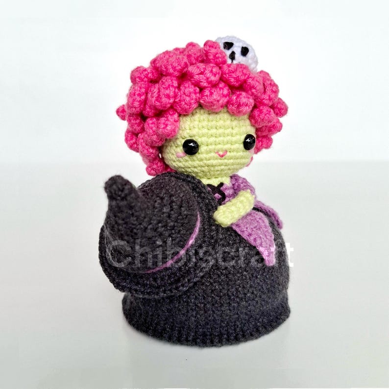 May include: A handmade crochet doll of a witch with pink curly hair, a green face, and a black dress. The doll has a small white skull on top of its head and a black witch hat. The doll is wearing a purple scarf.