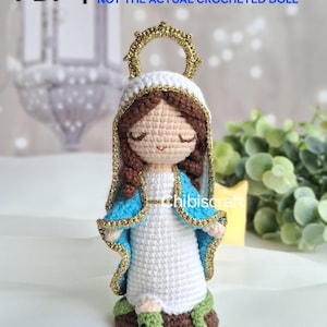 Puede incluir: Muñeca de ganchillo de una figura religiosa, con vestido blanco, chal azul y detalles dorados. La muñeca tiene cabello castaño y un halo dorado. La base es marrón con una serpiente verde. El texto en la imagen dice "PDF crochet pattern NOT THE ACTUAL CROCHETED DOLL".