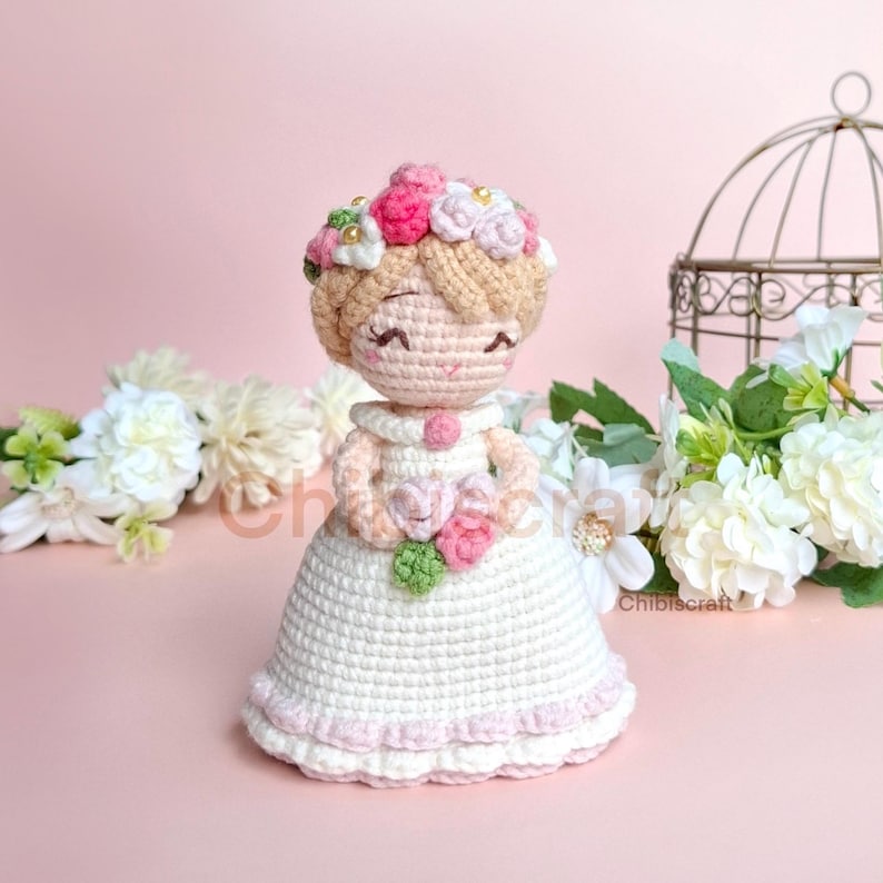 以下が含まれることがあります： 白いドレスにピンクのアクセントと花冠をつけた、かぎ針編みの人形。人形は、ピンクと白の花束を手に持っています。