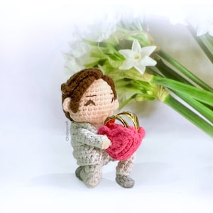 Op de afbeelding: Een gehaakt amigurumi-figuur van een persoon die een roze hartvormige doos vasthoudt met gouden ringen erin. Het figuur draagt ​​een grijs pak en heeft bruin haar. De achtergrond is een wit oppervlak met een boeket witte bloemen.