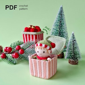 May include: A crocheted elf doll pops out of a striped gift box with a red bow. The box has a green lid that says "push!" and is sitting on a green surface with two small artificial Christmas trees.