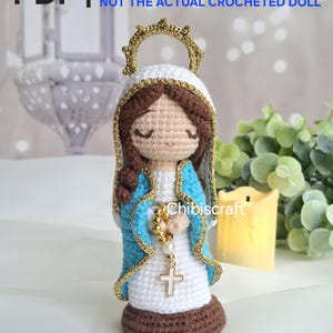 Op de afbeelding: Gehaakte pop van de Maagd Maria, met een witte jurk, blauwe sjaal en bruin haar. De pop heeft een gouden halo en houdt een rozenkrans met een kruis vast. De afbeelding bevat de tekst "PDF | crochet pattern".