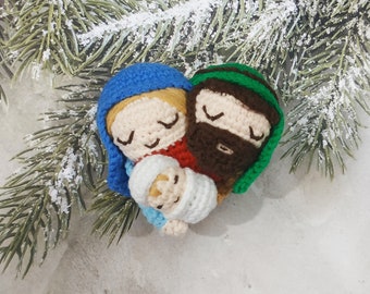 May include: Crocheted figures of a family, a woman in blue, a man in green, and a baby in white, with a snowy pine branch in the background.