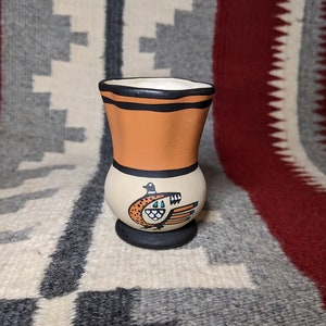 May include: A small, hand-painted ceramic vase with a black rim and a brown, orange, and white design. The vase features a stylized bird with black, white, and blue details.