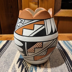 May include: A white ceramic vase with a geometric pattern in black, orange, and brown. The vase has a scalloped rim and a rounded body.