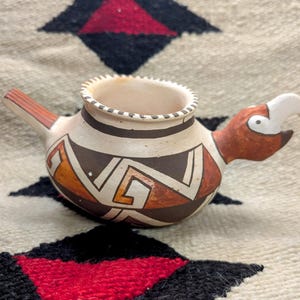 May include: A small, hand-painted ceramic bird-shaped pot with a geometric pattern in brown, orange, and white. The pot has a spout and a bird's head with a white beak.
