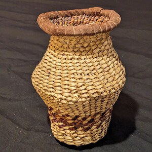 May include: A woven basket with a brown leather rim. The basket is made of natural materials and has a light brown color. The basket is a traditional style and is perfect for displaying flowers or other decorative items.