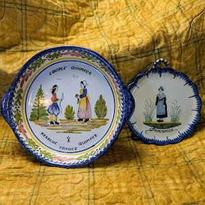 May include: Two ceramic plates with hand-painted designs. One is a large, round serving dish with handles, featuring a couple in traditional attire and the text "Couple Quimper, France." The other is a smaller, scalloped plate with a woman in a dress and the word "Provence."