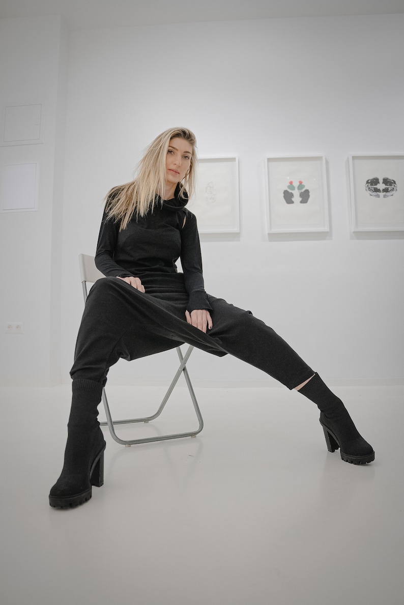 Peut inclure: Une femme portant une combinaison noire &agrave; manches longues est assise sur une chaise pliante argent&eacute;e devant un mur blanc avec des &oelig;uvres d'art encadr&eacute;es.