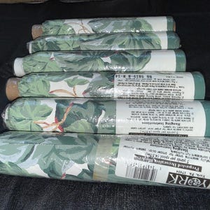 May include: Six rolls of prepasted decorative wallpaper border featuring a green and white botanical leaf pattern. Each roll is wrapped in clear plastic and includes a white label with black text detailing hanging instructions and product info.