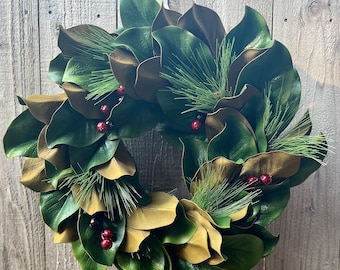 May include: A circular wreath composed of large, glossy green and brown magnolia leaves. Sprigs of green pine needles and clusters of small red berries are interspersed throughout the wreath, creating a festive, natural look. The wreath is displayed against a wooden background.