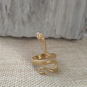 Op de afbeelding: Een goudkleurige slangenring met een textuuroppervlak. De kop van de slang is omhoog en het lichaam kronkelt. De ring staat tegen een neutrale achtergrond, wat het ontwerp van de sieraden benadrukt.