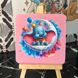 Peut inclure: Un dessous de verre carré rose avec une illustration de Dumbo l'éléphant sur un croissant de lune. Le motif comprend un chapiteau de cirque, un ballon bleu et des accents colorés. Le dessous de verre est présenté sur un petit chevalet en bois.