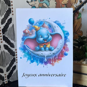 Peut inclure: Carte de vœux avec une illustration à l'aquarelle de Dumbo l'éléphant assis sur un croissant de lune. La carte est bleue, rose et jaune, avec le texte français "Joyeux anniversaire" en bas.