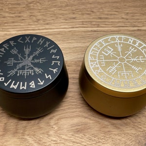 Könnte beinhalten: Zwei runde Metallbehälter mit Deckeln. Der Deckel des Behälters links ist schwarz mit einem silbernen Kompassdesign und Runen am Rand. Der Deckel des Behälters rechts ist golden mit einem weißen Kompassdesign und Runen am Rand.