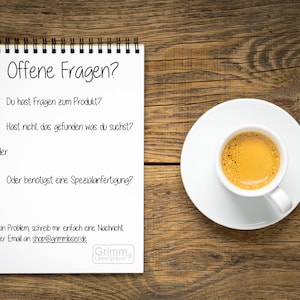 May include: A white notepad with a black spiral binding on a wooden surface. The notepad has the text "Offene Fragen?" at the top and a list of questions in German. The text at the bottom of the notepad reads "Kein Problem, schreib mir einfach eine Nachricht oder Email an shop@grimmlaser.de" and "Grimm Lasergravur". A white saucer with a white cup of coffee is on the right side of the image.