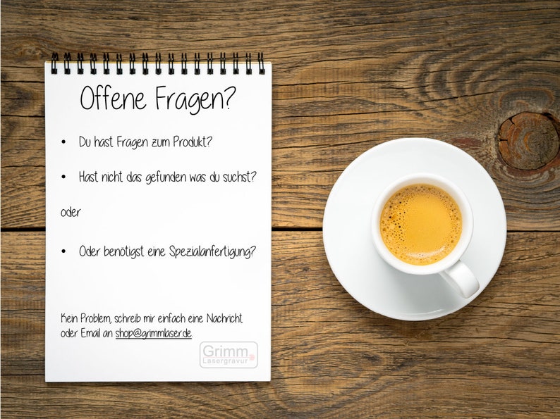 May include: A white notepad with a black spiral binding on a wooden surface. The notepad has the text "Offene Fragen?" at the top and a list of questions in German. The text at the bottom of the notepad reads "Kein Problem, schreib mir einfach eine Nachricht oder Email an shop@grimmlaser.de" and "Grimm Lasergravur". A white saucer with a white cup of coffee is on the right side of the image.